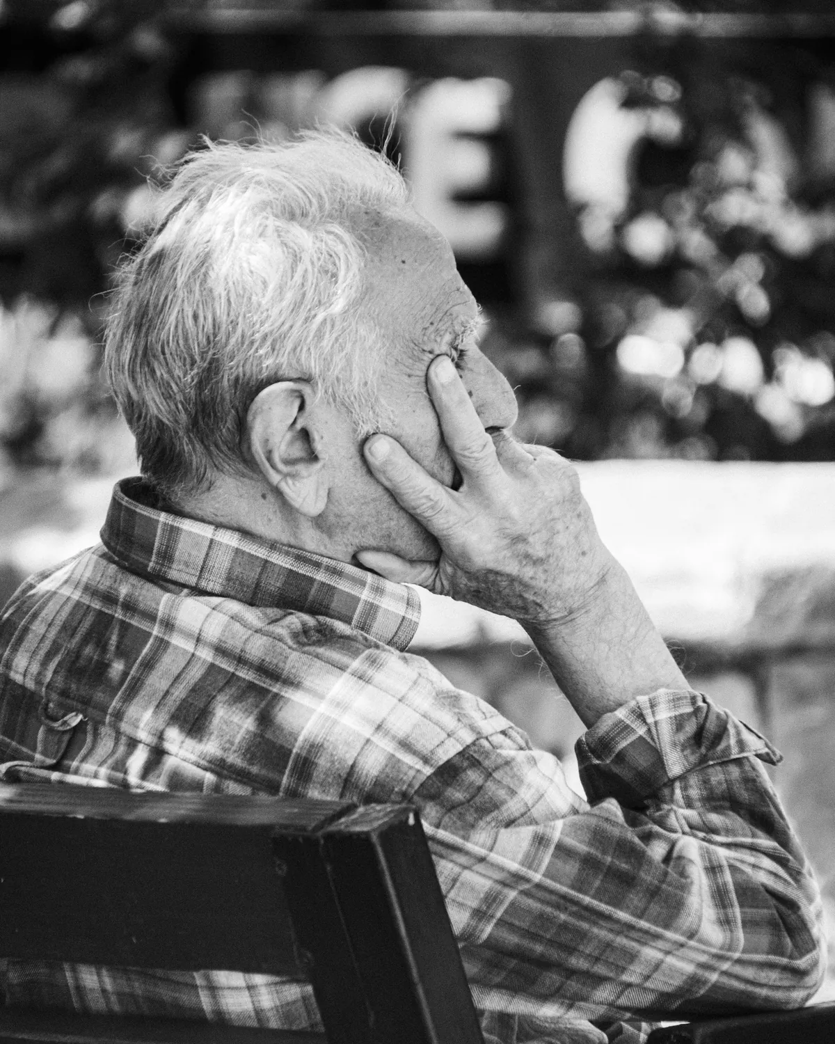 An older man sits on a bench with his hand against his cheek, looking ahead.